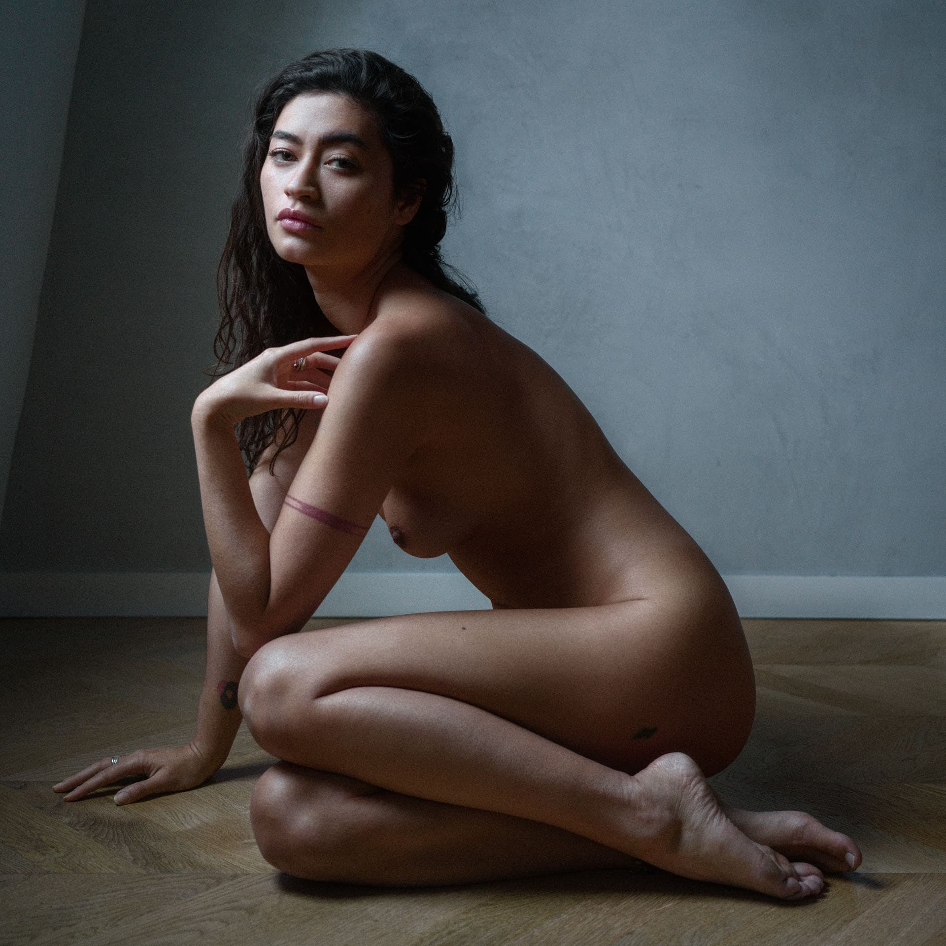 Jeune femme assise au sol en studio, regard tourné vers l'objectif, lumière douce sur parquet en chevrons.