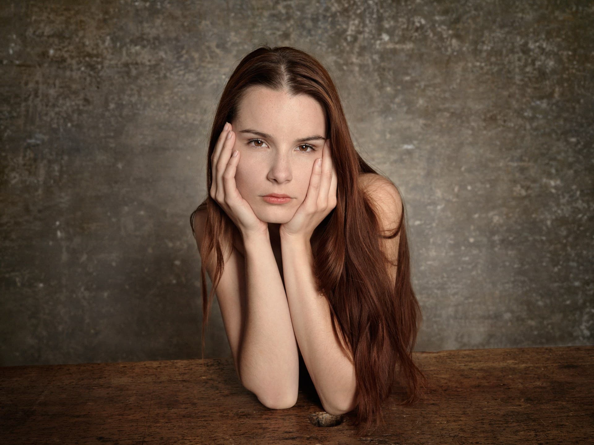 Face cradled in her hands, long auburn hair against a weathered wall.